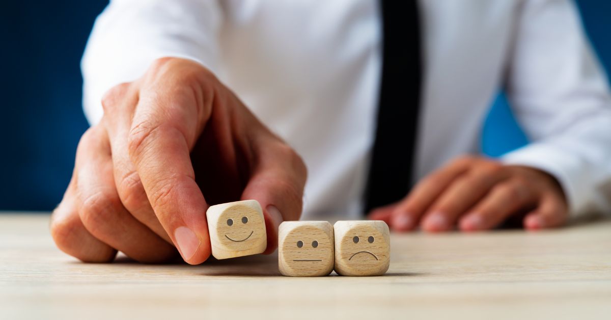 Close na mão de um homem de camisa e gravata mexendo em três dados que estão alinhados horizontalmente. Os dados são de madeira e têm rostinhos desenhados, o primeiro é feliz e os outros tristes.