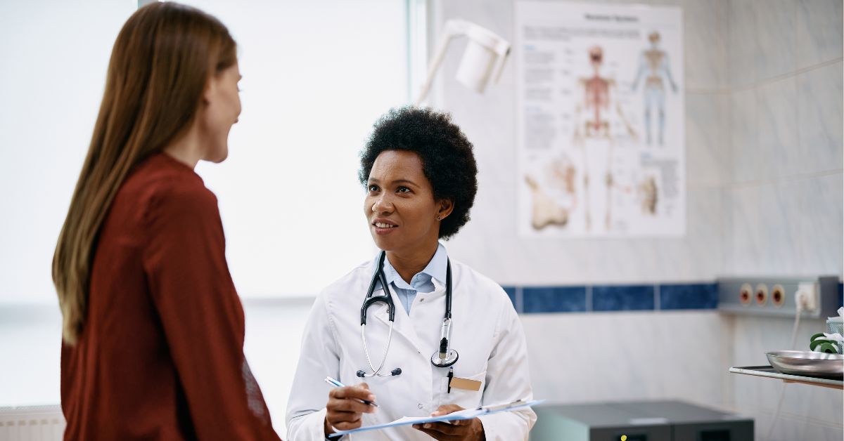 Médica negra sentada atendendo uma paciente mulher, que está de costas e em pé.