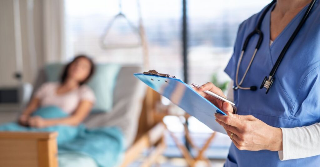 Médica, em primeiro plano, com prancheta na mão, e pessoal deitada em uma cama de hospital ao fundo.
