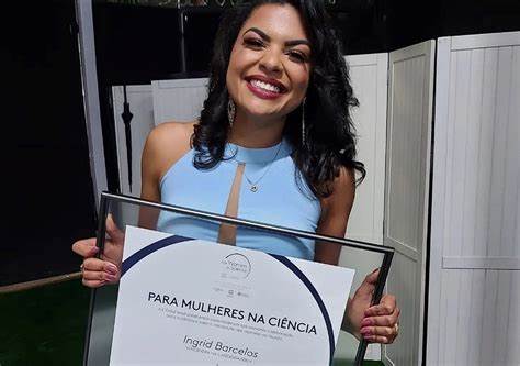 Mulher de cabelos soltos cacheados, vestindo uma roupa azul, sem mangas e segurando a placa do prêmio L'Oréal Para Mulheres na Ciência.