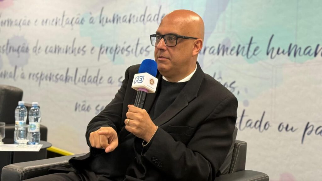 Homem de meia-idade, careca de terno escuro, sentado, falando ao microfone.