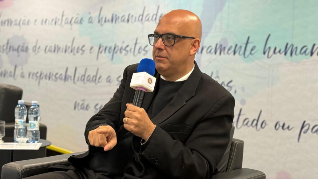 Luciano Sathler, um homem branco, careca, com óculos de aros pretos. Sentado em uma poltrona, com microfone na mão dando palestra. Ele usa um terno preto.